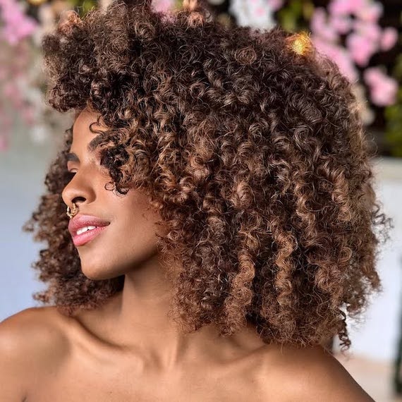 Model with chocolate brown, shoulder-length, curly hair, featuring caramel and blush balayage.