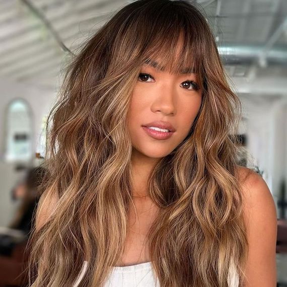 Model with long, light brown, sun-kissed hair, featuring tousled waves and blunt bangs.