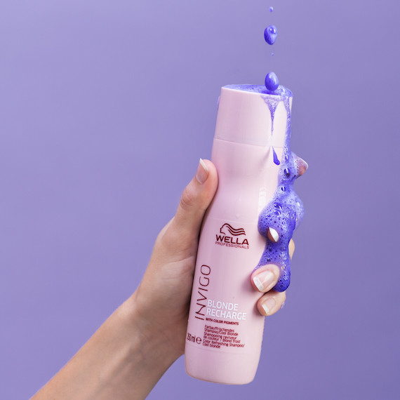 Model holds a bottle of INVIGO Blonde Recharge Shampoo, with the purple pigment pouring out. 