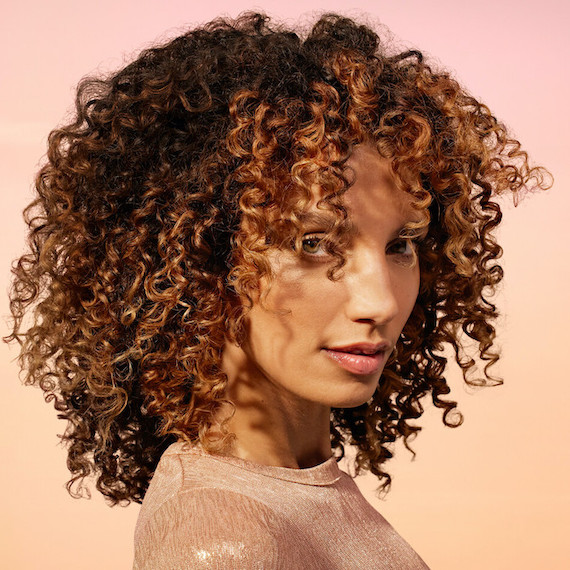 Model with dark brown, curly hair featuring glossy caramel highlights.