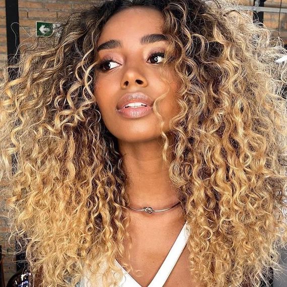 Model with curly hair that’s dark at the roots and bright honey blonde through lengths and ends.