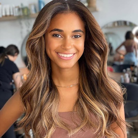 Model with long, dark brown, loosely curled hair, featuring glossy caramel balayage.