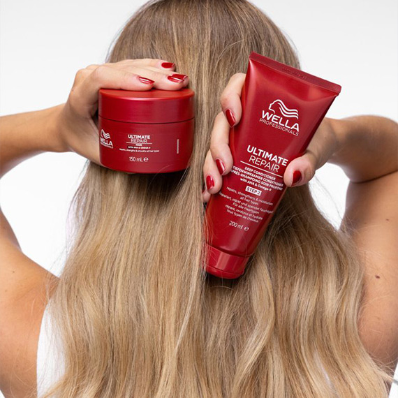 Model holds ULTIMATE REPAIR Mask and ULTIMATE REPAIR Conditioner in front of their blonde hair.