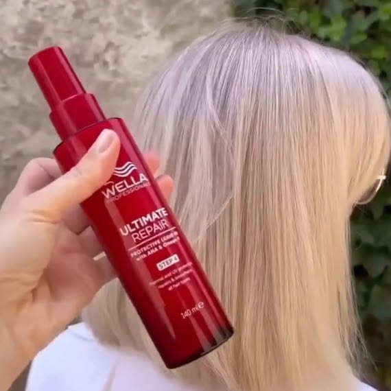 A bottle of ULTIMATE REPAIR Protective Leave-In is held up in front of a model’s blonde bob haircut.