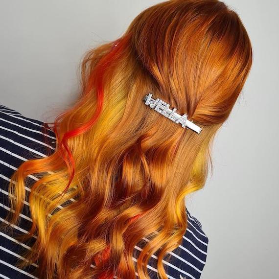 Back of woman’s head with long, wavy, sunset-colored hair in a half-up style with a ‘WELLA’ clip.