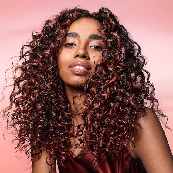 Model with long, dark brown, curly hair featuring coral pink highlights.