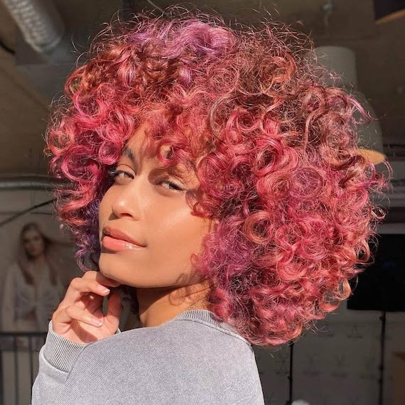 Model with full, curly hair featuring watermelon pink and purple highlights.