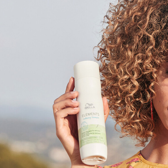 A model with curly hair holds up a bottle of Elements Calming Shampoo. 