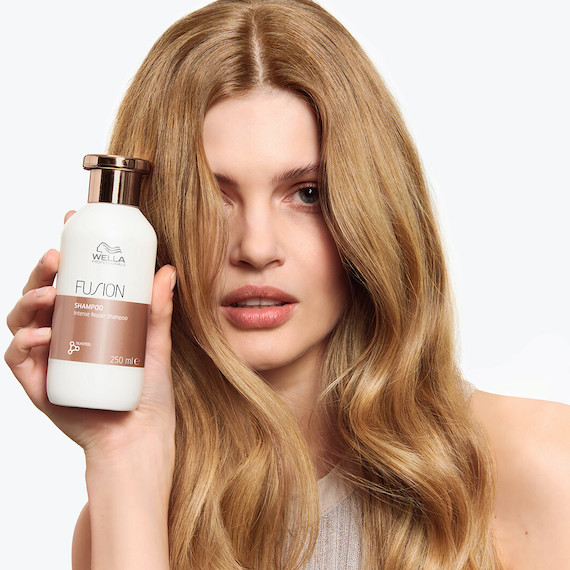 Model with golden blonde hair holds up a bottle of Fusion Intense Repair Shampoo.