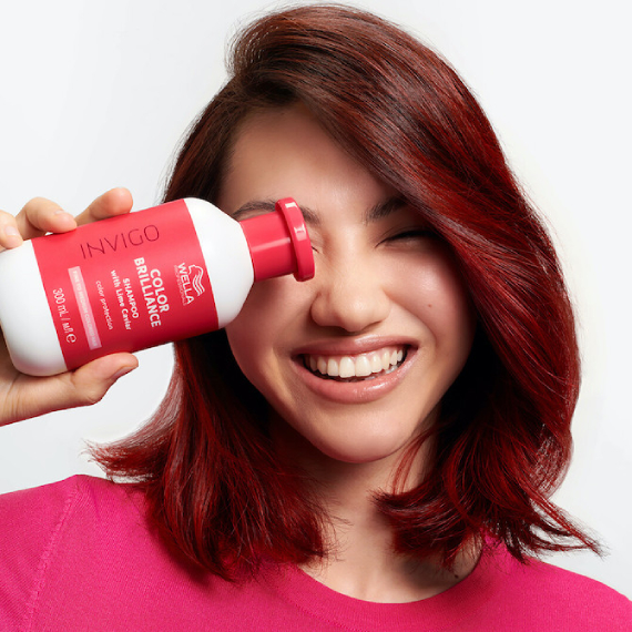 Model with dark red, shoulder-length hair holds up a bottle of INVIGO Color Brilliance Color Protection Shampoo.
