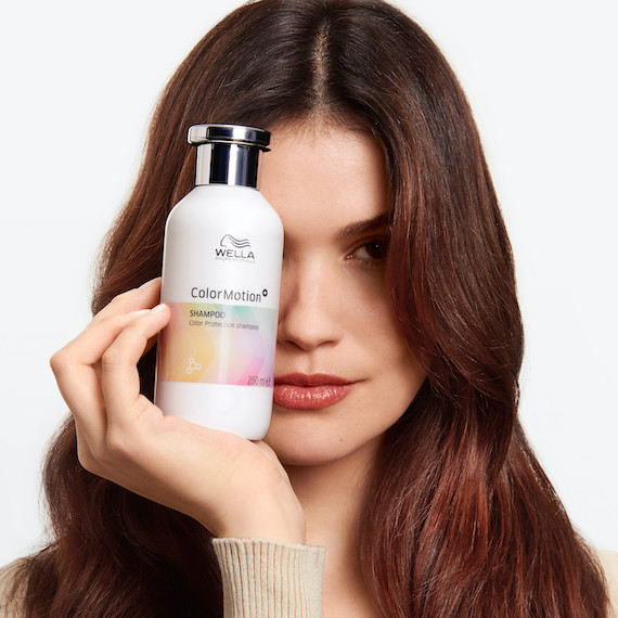 Model with long, dark brown hair holds up a bottle of ColorMotion+ Color Protection Shampoo.