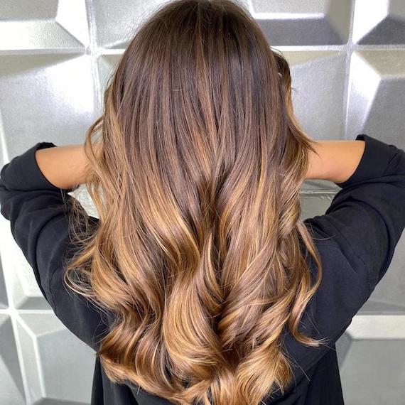 Back of woman’s head with long, loosely curled brown hair and toffee balayage.