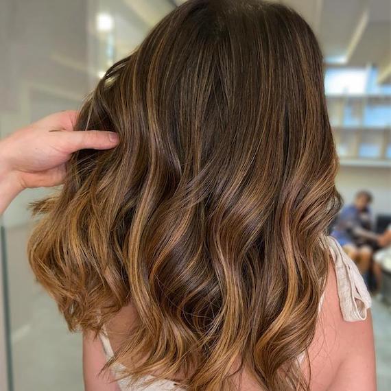 Back of woman’s head with long, wavy chocolate brown hair and golden twilights, created using Wella Professionals. 