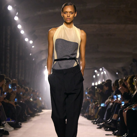 A model wearing a silk white and grey top with black trousers walks the runway.