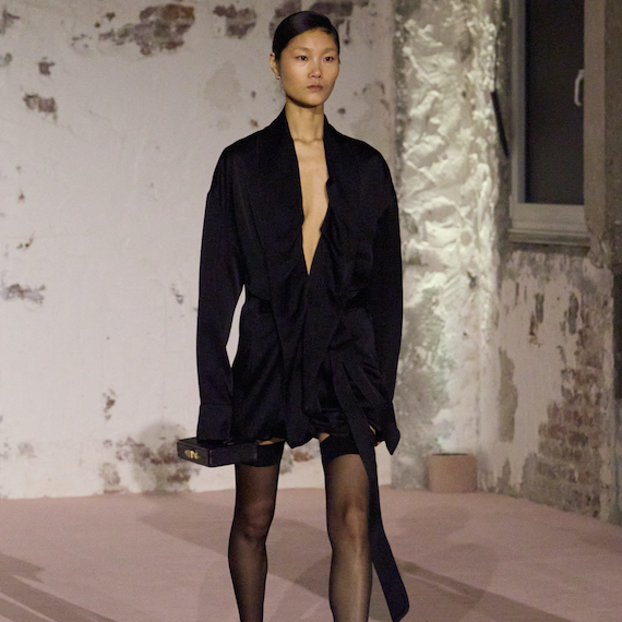 A model wearing a black silk shirt dress and thigh-high black stockings walks the runway.