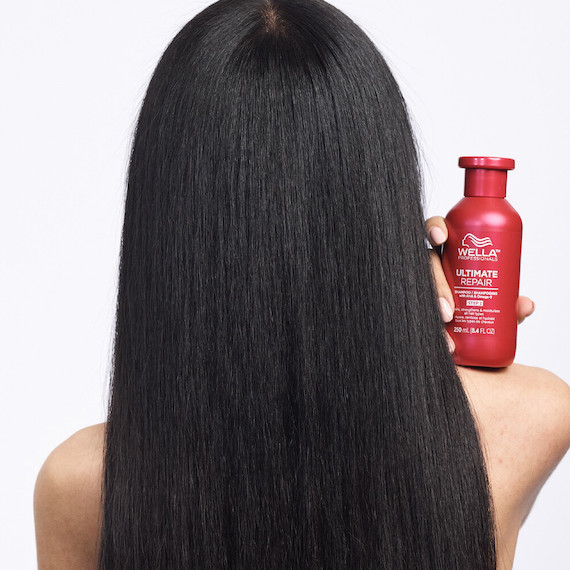 Ein Model mit langen, schwarzen Haaren steht mit dem Rücken zur Kamera und hält eine Flasche Wella Ultimate Repair Shampoo auf der Schulter