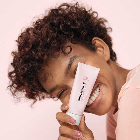 Model with short, brown, curly hair holding up a tube of SHINEFINITY.