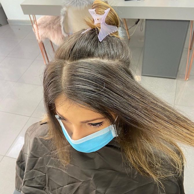 Woman with long dark hair and gray roots, sitting in a salon chair.