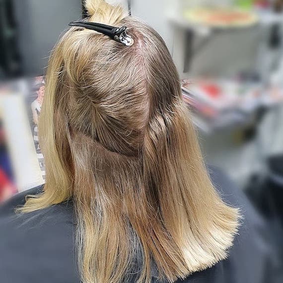 Back of woman’s head with shoulder-length blonde hair and gray roots, sitting in a salon chair. 