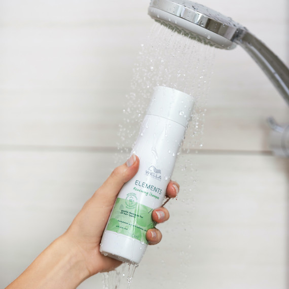 A hand holds Elements Renewing Shampoo underneath the showerhead. 