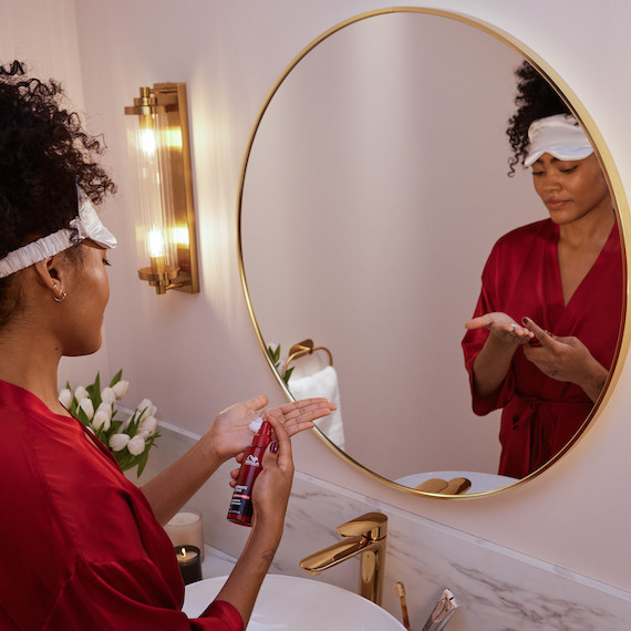 A model with dark, curly hair stands in front of the bathroom mirror and applies ULTIMATE REPAIR Night Serum. 