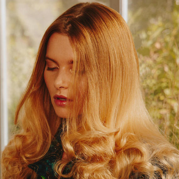 Model gazing downwards. Her golden blonde hair is styled in loose, lightly-tousled waves.