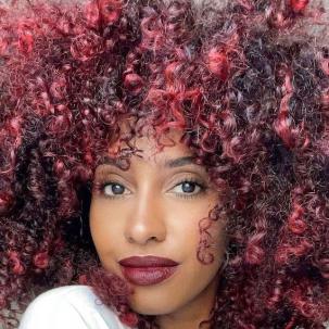 Headshot of a person with voluminous, black and red curly hair. They wear red lipstick to match.