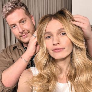 Hairdresser styles model’s blonde, wavy hair while they both look into the camera.