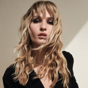 Model with sandy blonde, wavy hair and a fringe faces the camera.