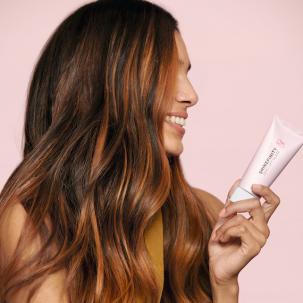 Woman with long, brown glossy hair smiling while holding a tube of the Shinefinity Glaze.