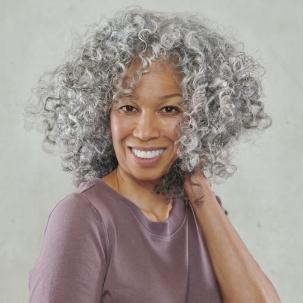 Woman facing the camera and smiling as she clutches her grey, curly hair. 