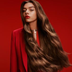 Model mit sehr langem, braunem, gewelltem Haar, das nach vorne schaut und einen roten Blazer und roten Lippenstift trägt