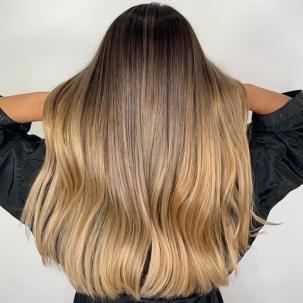 A person with long blonde ombre hair wearing a black robe faces a light grey wall