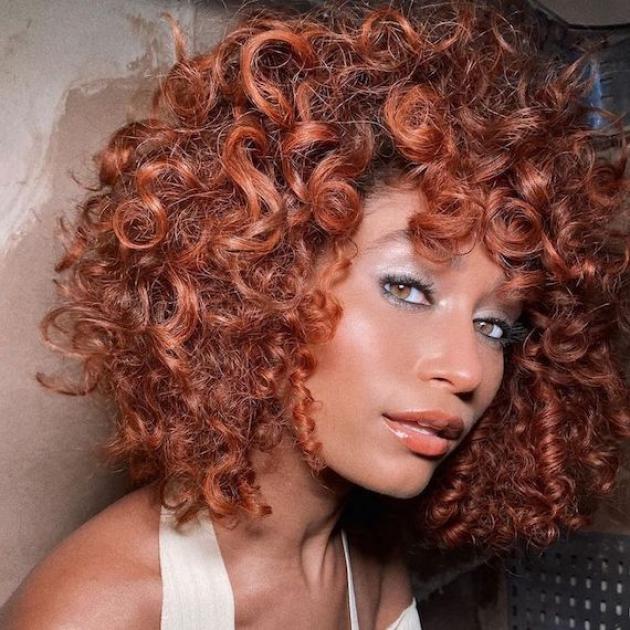 Model will full, voluminous, curly, copper red hair looks directly into the camera.