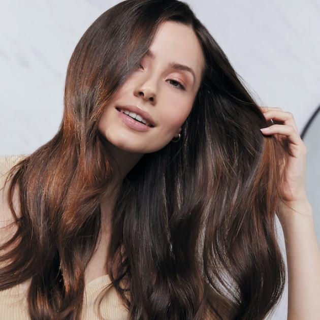 Model runs hands through long, glossy, brown hair while smiling at camera.