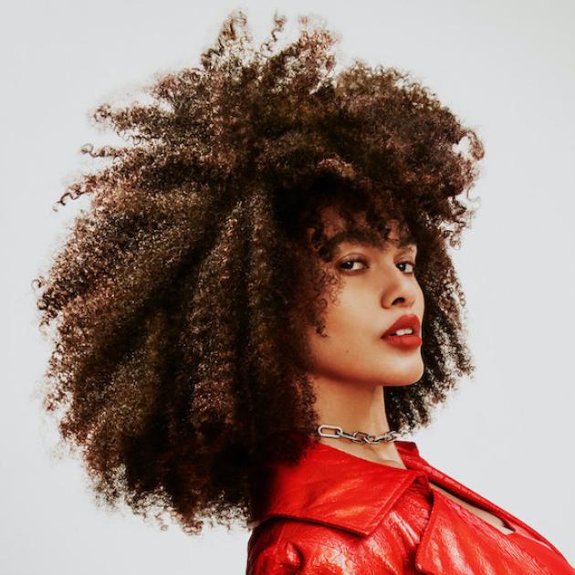 Headshot of a model with very curly brown hair wearing a red jacket and red lipstick