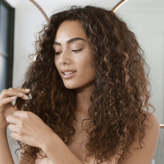 Person with long, brown curly hair holds a section of their curls and looks at them