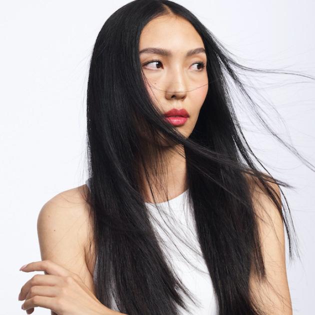 A model with long dark hair wearing red lipstick. They look to the side with their hair blowing in the same direction