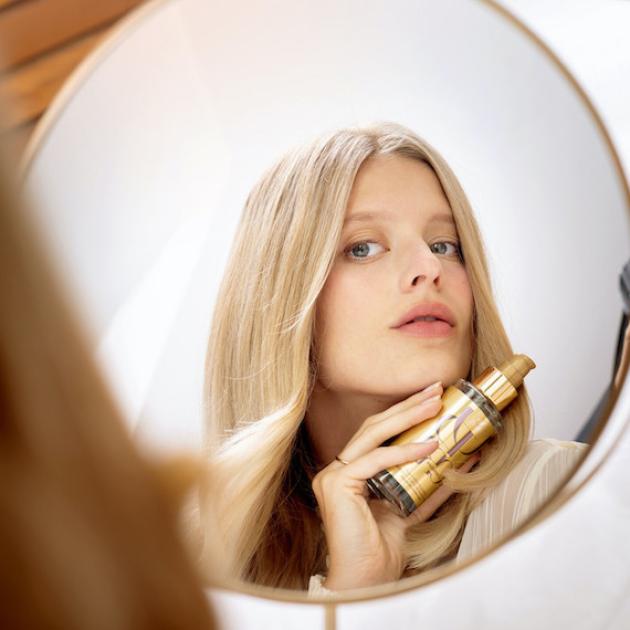 Frau mit langem blondem Haar schaut in den Spiegel, während sie eine Flasche Oil Reflections Öl von Wella Professionals hält. 